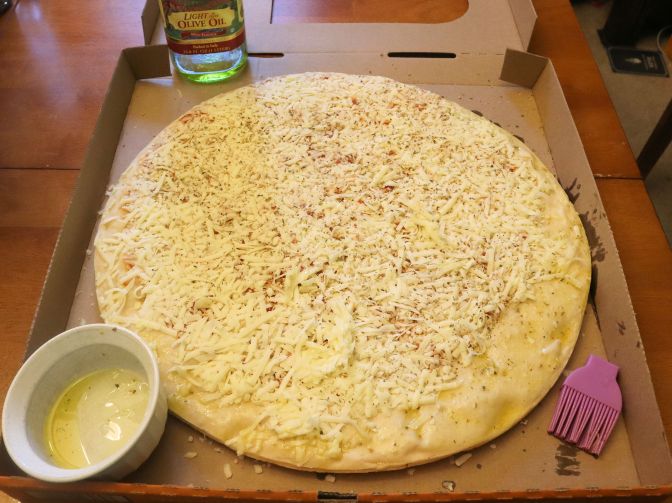 Picture of uncooked pizza with bottle of olive oil, ramekin with a small amount of olive oil in it, as well as the head of a purple silicone food brush.  (Who knows where the handle went!) 